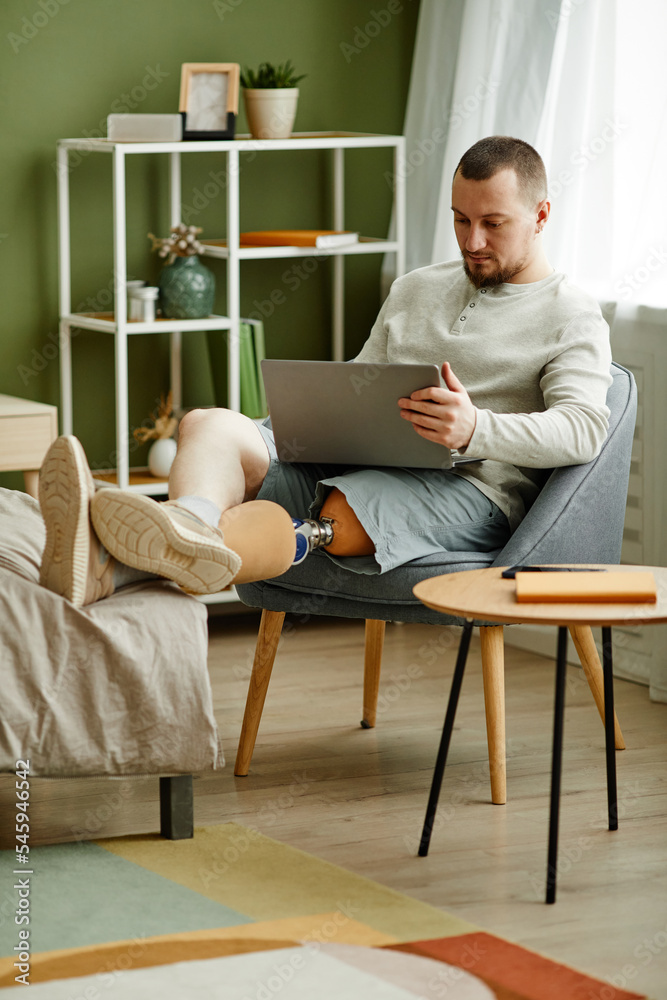 Vertical full length portrait of adult man with prosthetic leg relaxing at home and using laptop Vertical full length portrait of adult man with prosthetic leg relaxing at home and using laptop