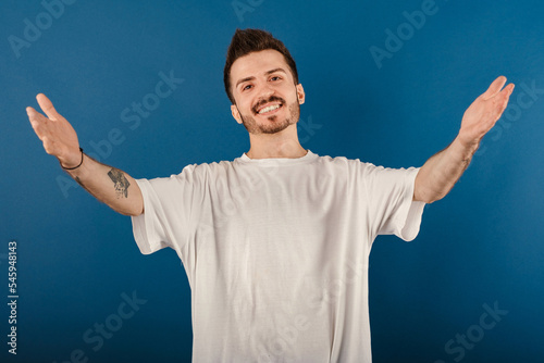 Happy young man wearing white t-shirt posing isolated over blue background smiling with open arms for hug. Cheerful expression embracing happiness.