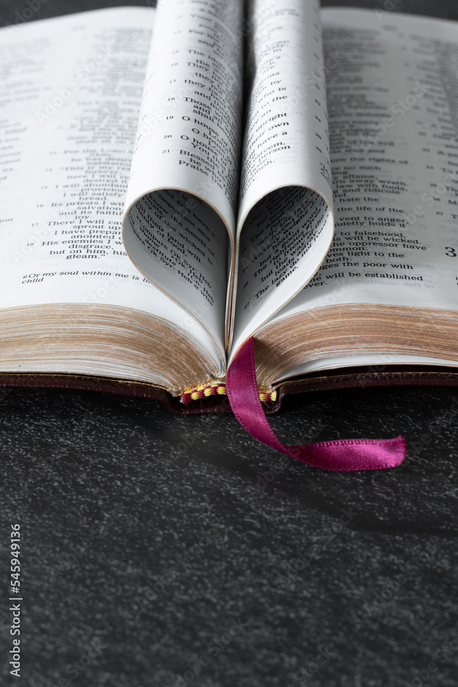Fototapeta premium Open Holy Bible Book with golden pages folded in a heart shape on dark background with copy space. Vertical shot. Christian biblical concept of love and faith.
