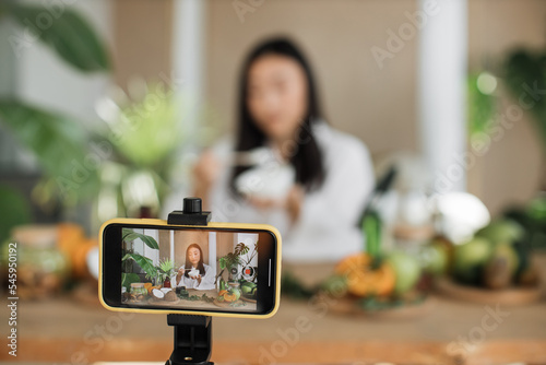 Young asian woman blogger or content creator in white bathrobe sitting at wooden table with various ingredients preparing natural cosmetics at home, mixing the mass in a bowl while preparing cream.