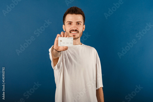 Handsome caucasian man wearing white t-shirt posing isolated over blue background showing his plastic credit or debit card with hands. Shopping and finance concept.