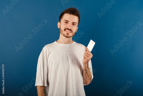 Handsome caucasian man wearing white t-shirt posing isolated over blue background holding plastic credit or debit card while looking at the camera.
