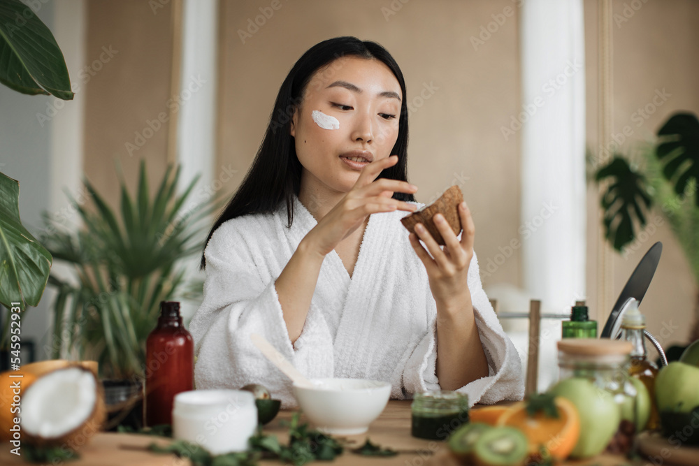Blogger or content creator asian woman preparing applying natural cosmetics at home, holding coconut for homemade cream for skin, hair care sitting at wooden table with exotic background in home spa Blogger or content creator asian woman preparing applying natural cosmetics at home, holding coconut for homemade cream for skin, hair care sitting at wooden table with exotic background in home spa
