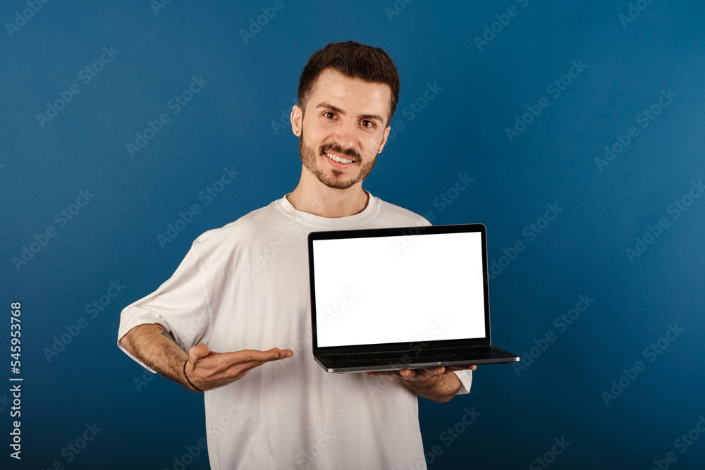 Handsome caucasian man wearing white t-shirt posing isolated over blue background holding laptop pc computer with blank empty screen.
