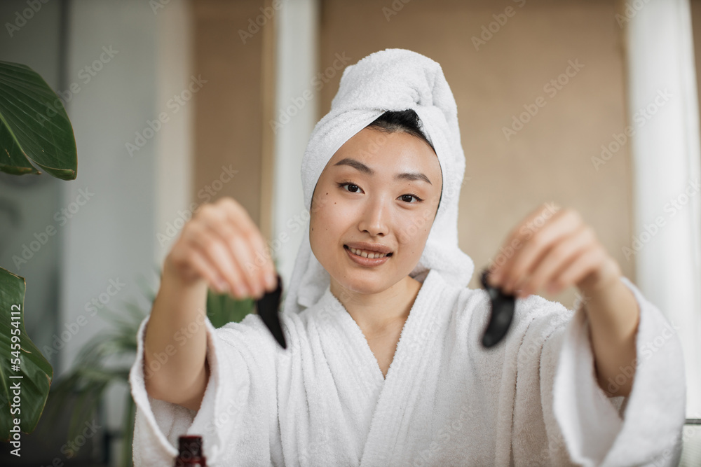 Pleasant asian woman in white bathrobe and towel holding eye patches for beauty procedures at exotic tropical living room. Beautiful female enjoying hygienic procedures. Pleasant asian woman in white bathrobe and towel holding eye patches for beauty procedures at exotic tropical living room. Beautiful female enjoying hygienic procedures.