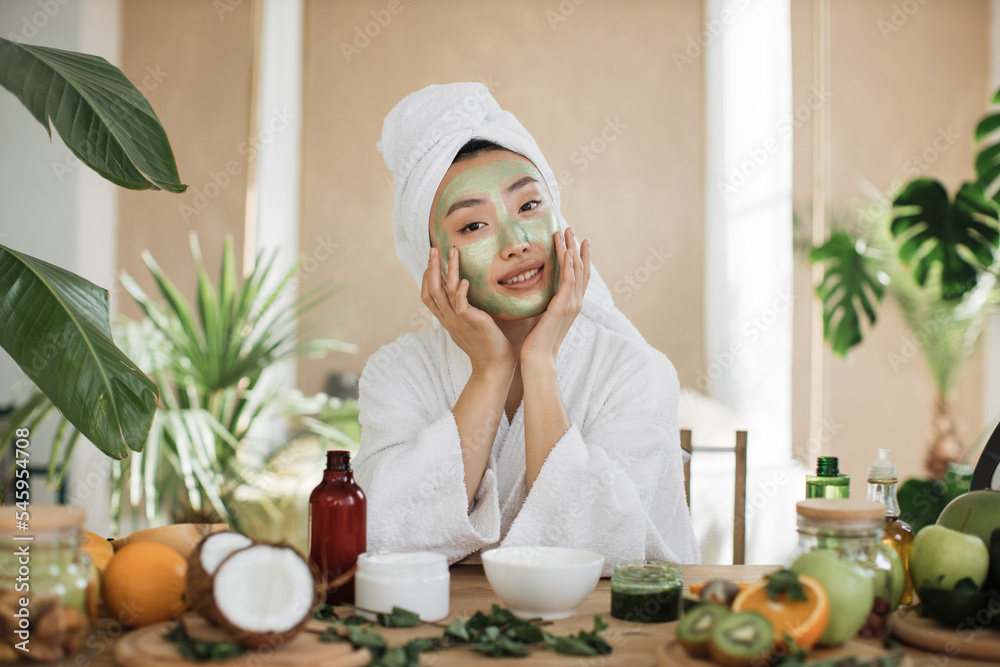 Portrait of beautiful young asian woman with green cosmetic mask on face and towel on head over exotic background. Pretty lady doing skin care procedures indoors at home. Portrait of beautiful young asian woman with green cosmetic mask on face and towel on head over exotic background. Pretty lady doing skin care procedures indoors at home.