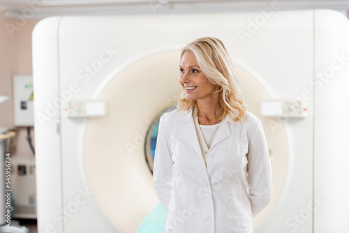 smiling blonde doctor in white coat looking away near ct scanner in hospital.