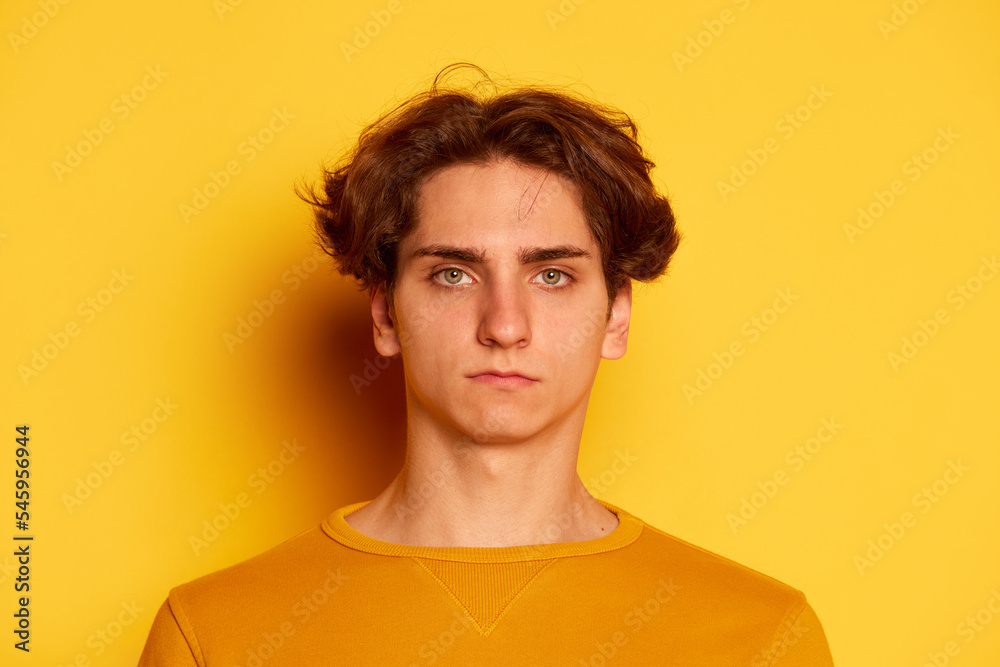 Portrait of young man with curly hair posing with serious face, looking at camera isolated over yellow background Portrait of young man with curly hair posing with serious face, looking at camera isolated over yellow background