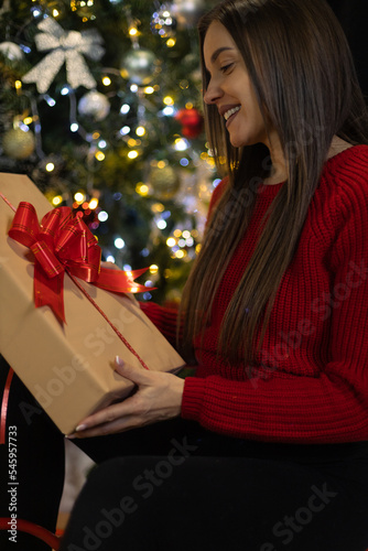 A smiling young girl, pregnant, with a smile on her face opens Christmas presents in front of the Christmas tree. The gifts are wrapped in decorative paper and tied with a red bow