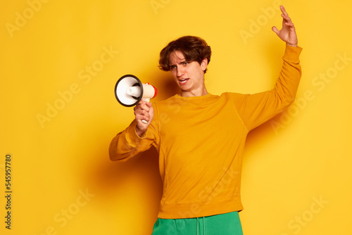 Portrait of young man with curly hair posing, emotively speaking in megaphone isolated over yellow background. News © master1305