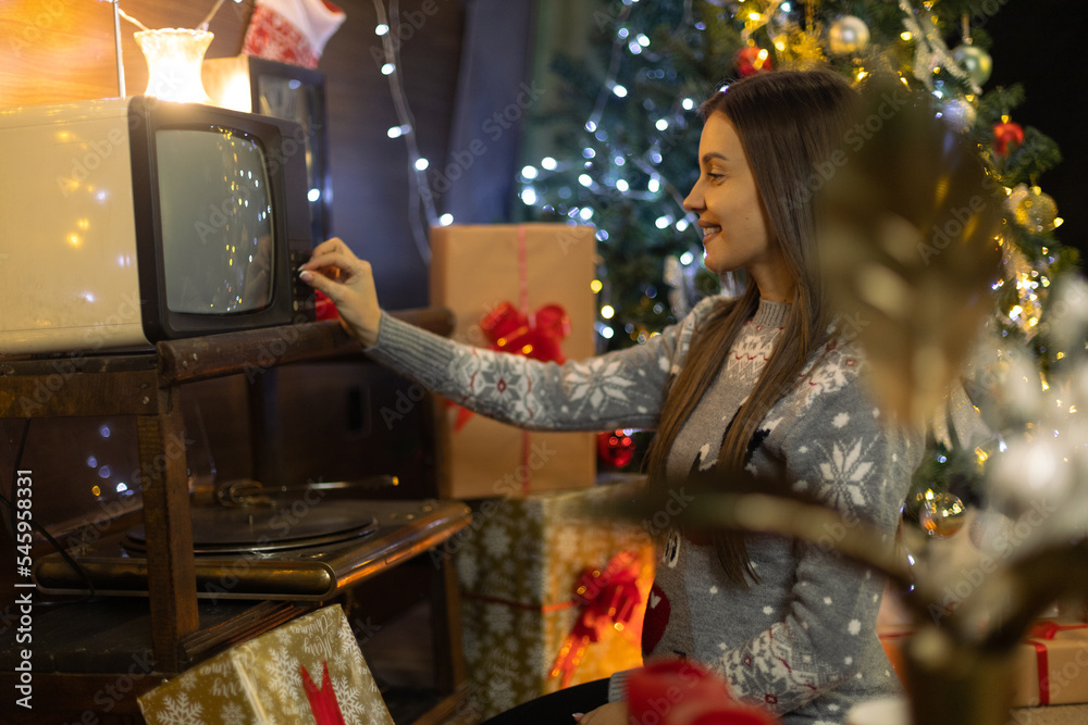 A smiling girl in front of a Christmas tree with presents is trying to turn on an old 1970s television and record player. The New Year's lighting scenography from the last century shines around it
