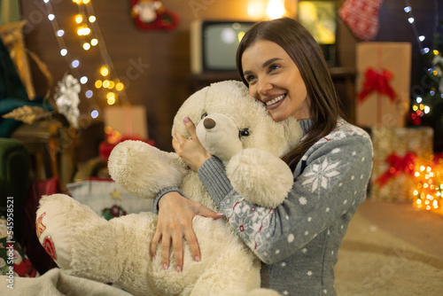Beautiful pregnant girl in front of Christmas tree and New Year decorations, sitting alone with a stuffed toy, a teddy bear, smiling, enjoying the background are gifts old TV and old furniture