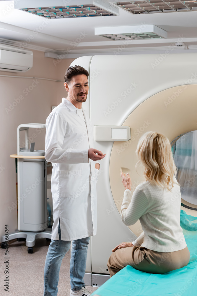 positive doctor in white coat talking to mature woman sitting near computed tomography scanner. positive doctor in white coat talking to mature woman sitting near computed tomography scanner.