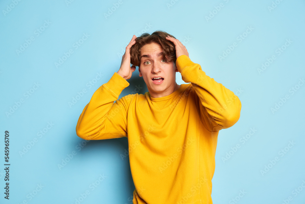 Portrait of young man with curly hair posing, holding head isolated over blue background. Shock and fear