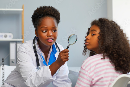 Concept of facial skin problems in adolescence. Female dermatologist examines teenage girl's face with magnifying glass for moles and redness. African American girl in dermatologist doctor's office.