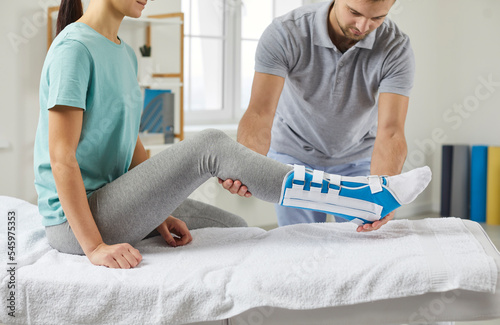 Woman with a splint on her shin lies on a couch in the physiotherapist's office in the rehabilitation center. Male physiotherapist examines a patient's leg during a rehabilitation consultation  photo