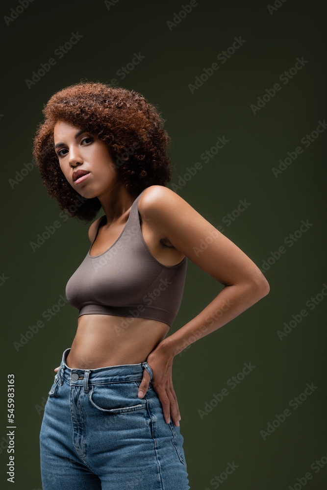 Curly african american woman in top and jeans holding hand on hip and looking at camera isolated on dark green. Curly african american woman in top and jeans holding hand on hip and looking at camera isolated on dark green.