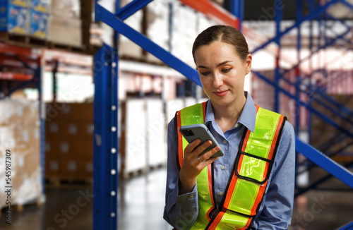 Female warehouse engineer wearing hard hat checking goods in warehouse report on smartphone