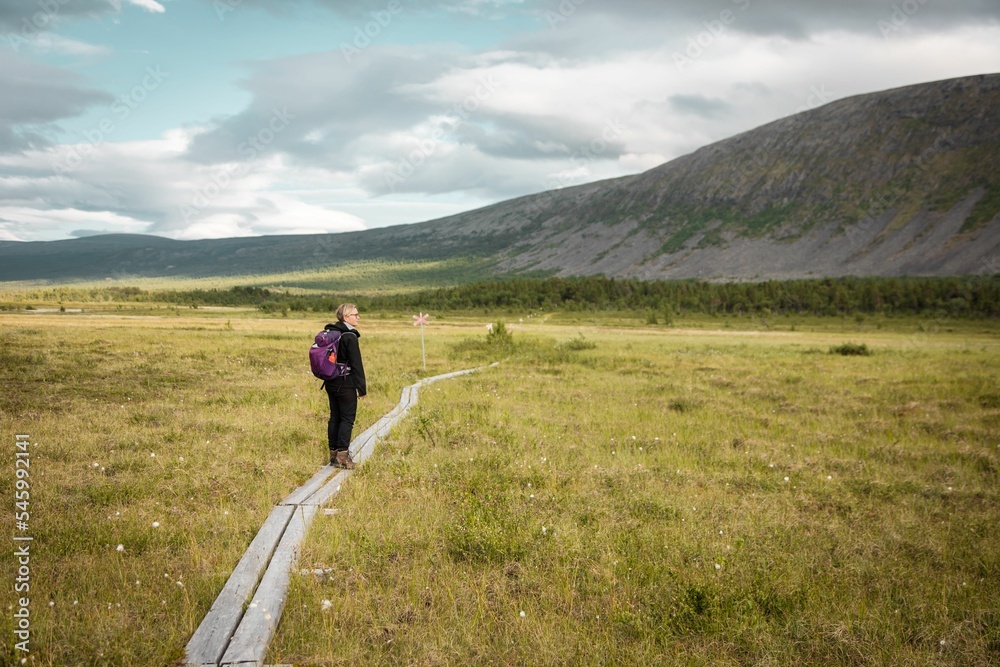Caucasian woman with backpack walking on a trail in a green field with mountain in the background Caucasian woman with backpack walking on a trail in a green field with mountain in the background