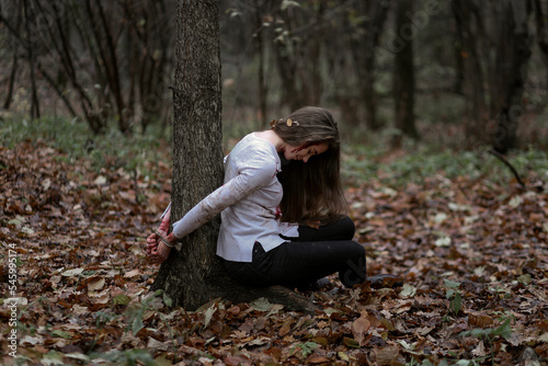 Fotografía Unconscious young woman tied to tree and sitting on ground in dark forest