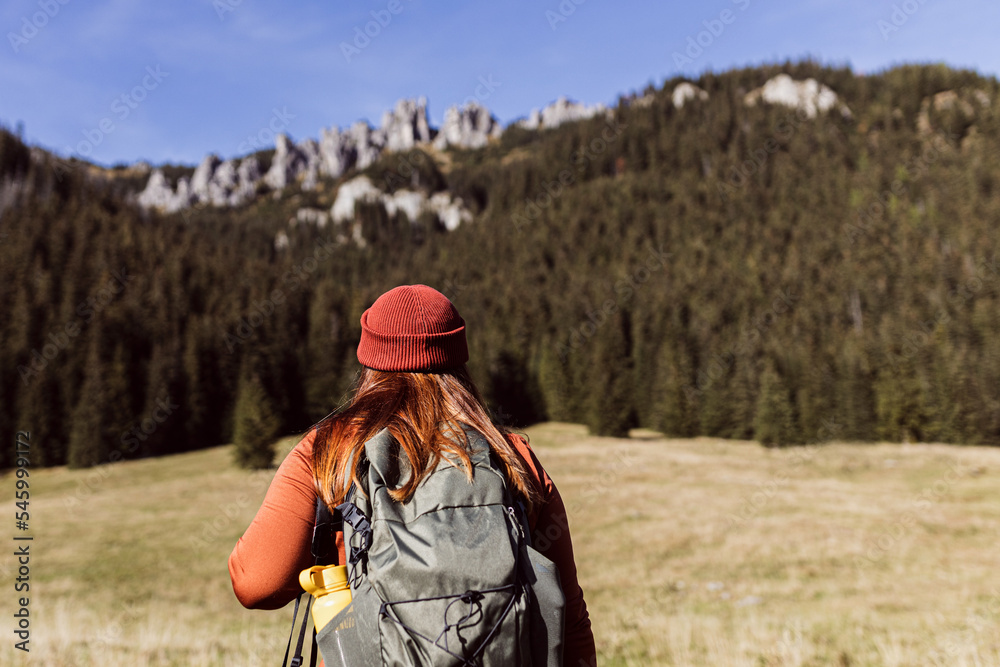 Back view of stylish woman wearing green backpack and red hat looking at mountain view while relaxing in nature. Travel and wanderlust concept. Amazing chill moment