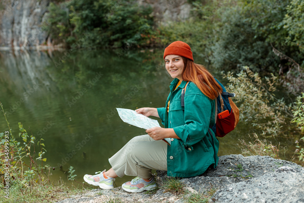 Stylish hipster woman holding paper map, wearing backpack and red hat relaxing in nature lake background. Travel and wanderlust concept. Amazing chill moment. Full body photo