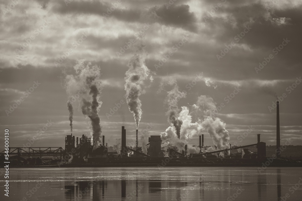 Grayscale of a smoking factory reflecting on a lake on a cloudy day in Hamilton, Ontario