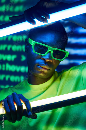 Cool trendy black man in sunglasses touching neon lamps in disco club photo