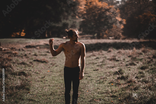 Man with a ponytail and one arm raised shirtless in a wooded area