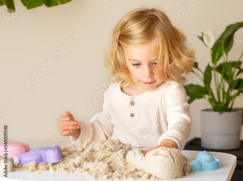 Cute happy caucasian,blonde,curly-haired toddler,baby girl playing with kinetic sand indoors.Preschool kid early development,motor,sensoric skills concept.Copy space.