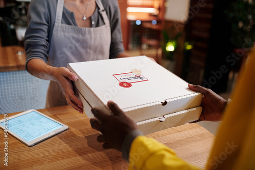 Hands of young female worker of cafe passing two packed square boxes of pizza to courier over wooden counter while preparing orders