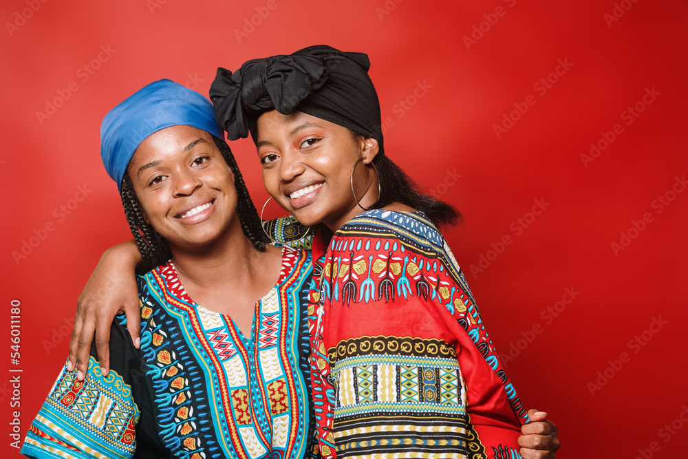African-American women laugh in embrace in a studio portrait on a red background. They are wearing a typical African dress very colorful.