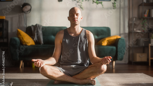 Fototapeta Naklejka Na Ścianę i Meble -  Athletic Young Man Exercising, Doing Meditation in the Morning in His Bright Sunny Room at Home. Handsome Bald Male Athlete in Sports Clothes Practising Mindfulness on Yoga Mat.