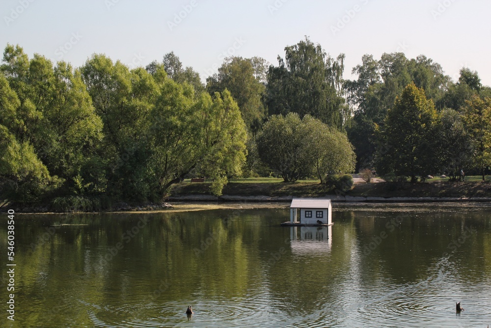 White wooden floating duck house in the centre of the pond in public park. Bird sanctuary in artificial lake. Spring nest. Summer green view. Wildlife nature reserve in river water. Waterfowl shelter White wooden floating duck house in the centre of the pond in public park. Bird sanctuary in artificial lake. Spring nest. Summer green view. Wildlife nature reserve in river water. Waterfowl shelter