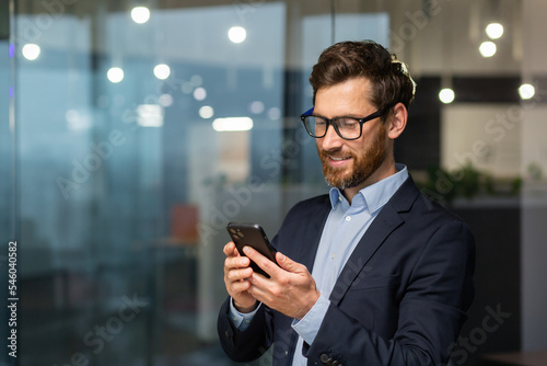 Successful financier investor works inside office at work, businessman in business suit uses telephone near window, man smiles and reads good news online from smartphone.