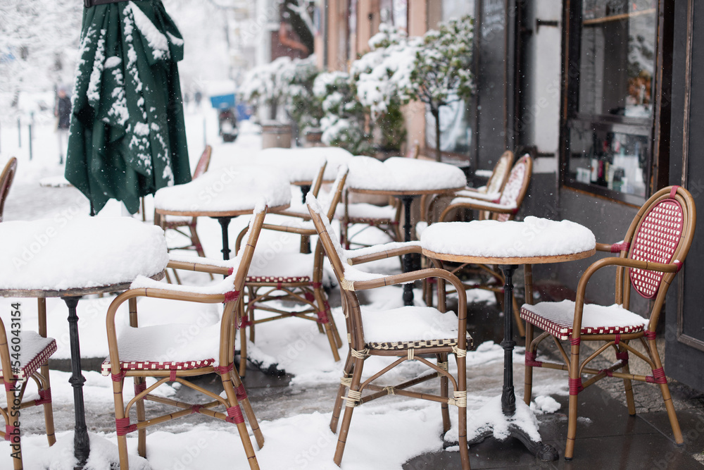 Snowy winter scene on beautiful city street, Cafe tables covered with snow in old European town, Outdoor restaurant terrace at cold snowy day