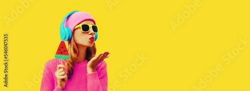 Summer fresh colorful portrait of stylish woman in headphones listening to music with fruit juicy lollipop or ice cream shaped slice of watermelon on yellow background