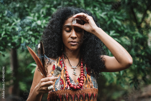 retrato de una mujer afroamericana al aire libre haciendo un ritual, con una mano sostiene algunas plumas y un rollo de hojas de salvia y con la otra coloca un cristal de cuarzo en su frente.  photo