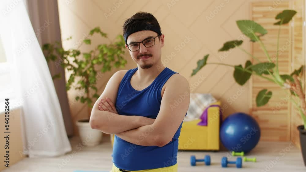 Nerd Chubby man with a moustache, glasses and headband in blue and yellow uniform nods and smiles looking at the camera. Fitness equipment visible at the background. Positive motivation