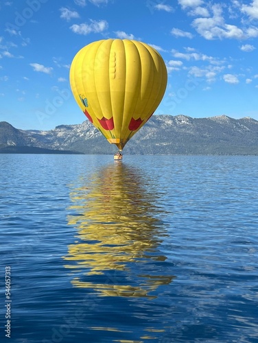 Vertical shot of a yellow hot air balloon flying over a tranquil lake against mountains
