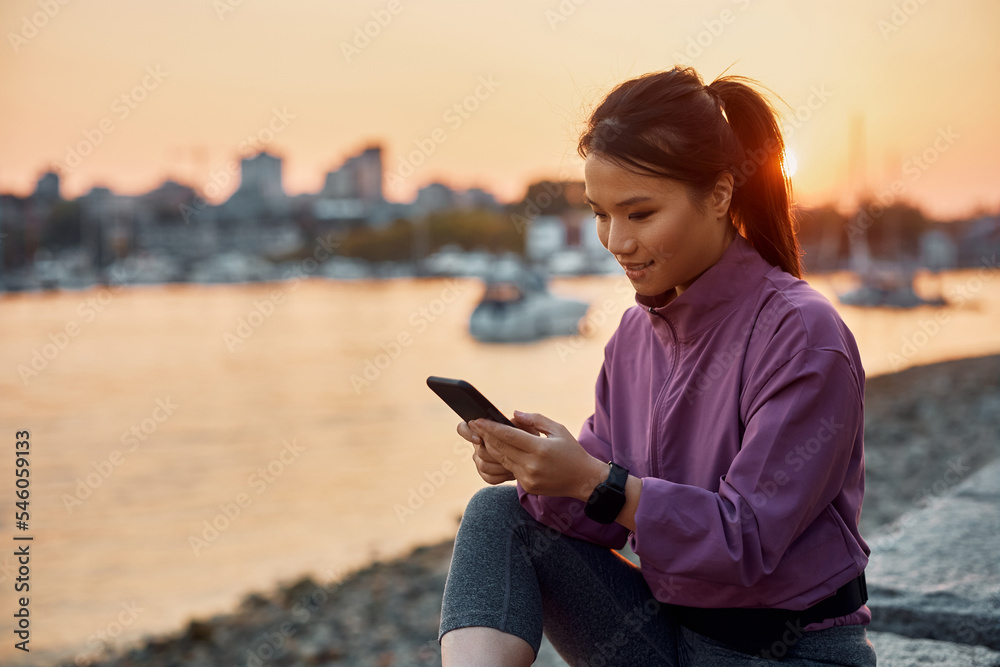 Smiling Asian athletic woman texting on cell phone at sunset.