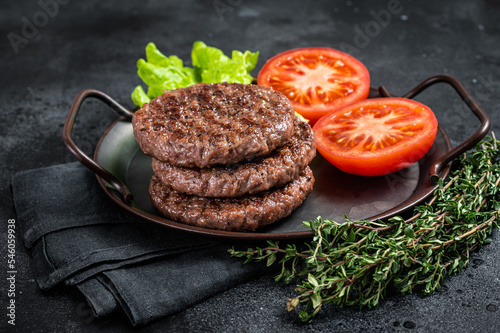 Tasty grilled burger beef patty with tomato, spices and lettuce in kitchen tray. Black background. Top view photo