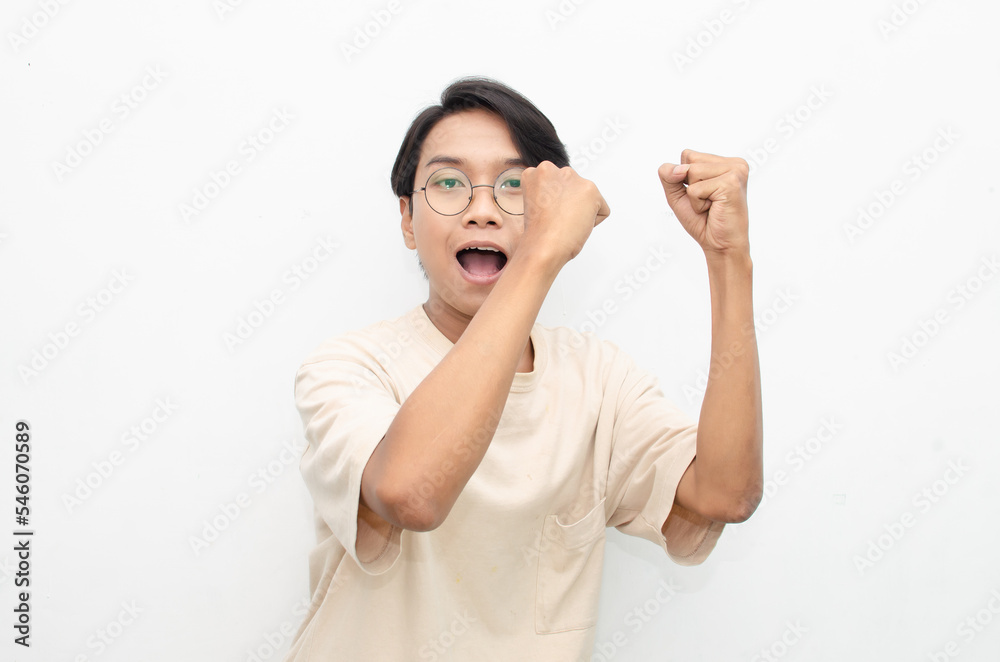 Asian young man wearing casual beige tshirt surprised, amazed, wow shocked raising arms gesturing victory and yes gesture Asian young man wearing casual beige tshirt surprised, amazed, wow shocked raising arms gesturing victory and yes gesture