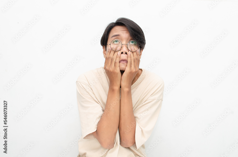 Young Asian man wearing glasses and beige tshirt with crying face expression isolated over white background. Crying face expression of Young Asian man with hand gesture. Young Asian man wearing glasses and beige tshirt with crying face expression isolated over white background. Crying face expression of Young Asian man with hand gesture.
