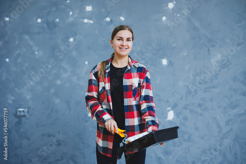 A beautiful young woman in a checkered shirt is renovating her room, holding a paint roller