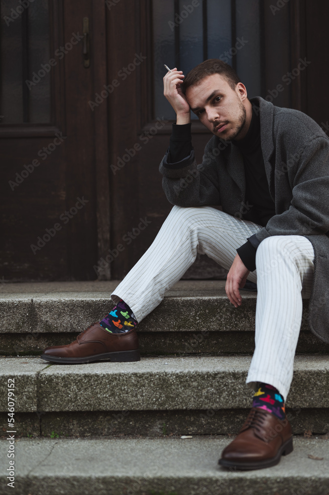 Young fashionable man sitting on stairs in the front of the building, looking thoughtful and smoking cigarette
