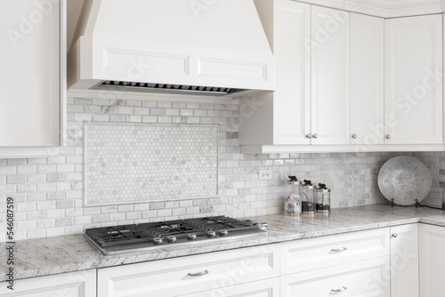 A kitchen stovetop and hood with off white cabinets, a tiled backsplash, and marble countertop. photo