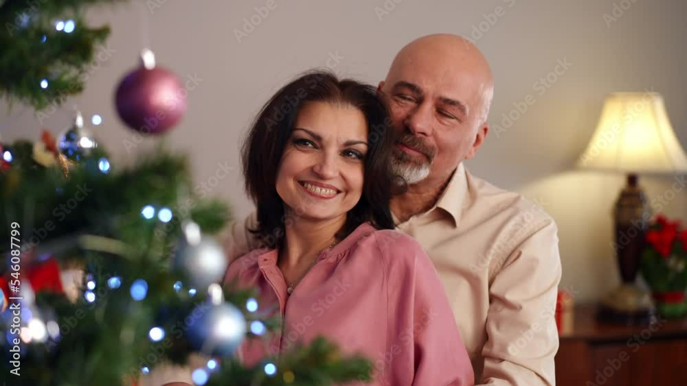Smiling Caucasian man and woman decorating Christmas tree at home hugging. Positive loving couple enjoying New Year preparations indoors embracing. Love and holiday celebration concept Smiling Caucasian man and woman decorating Christmas tree at home hugging. Positive loving couple enjoying New Year preparations indoors embracing. Love and holiday celebration concept