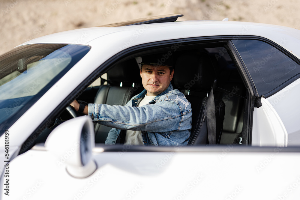 Handsome man in jeans jacket and cap sit at his white muscle car.