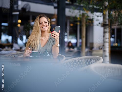 A happy woman sits in a cafeteria, texing messages and enjoying tea while smiling at the phone. photo